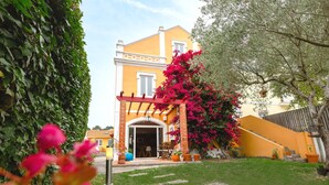 A yellow building with a covered entrance, surrounded by greenery and potted plants.