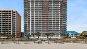 Et højt hotel med flere balkoner, en sandstrand i forgrunden og en klar blå himmel.