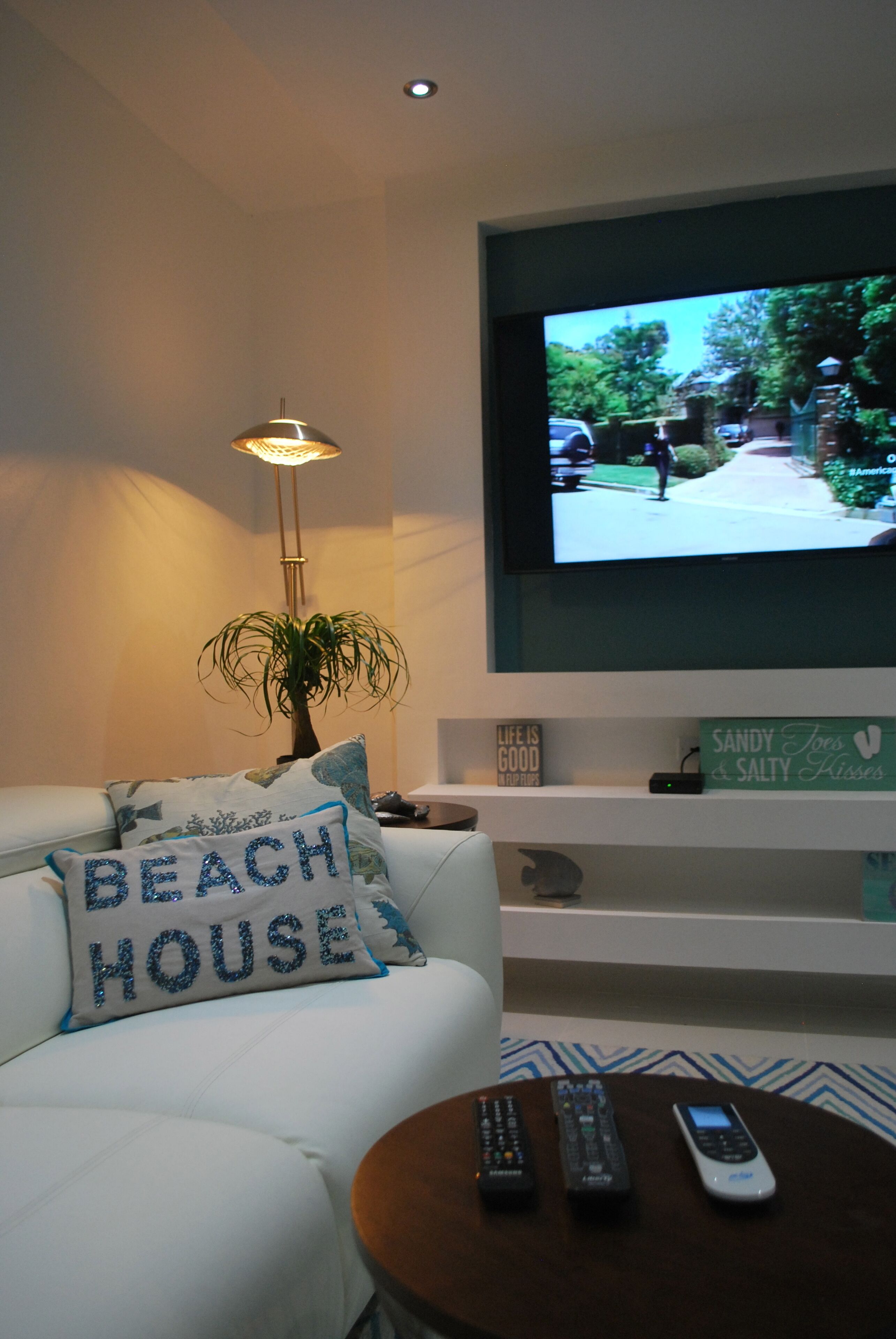Un salon avec un canapé blanc, une table basse avec des télécommandes, un téléviseur fixé au mur et une plante.