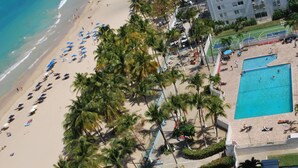 Luchtbeeld van een strand met palmbomen, een zwembad en een gebouw met balkons.