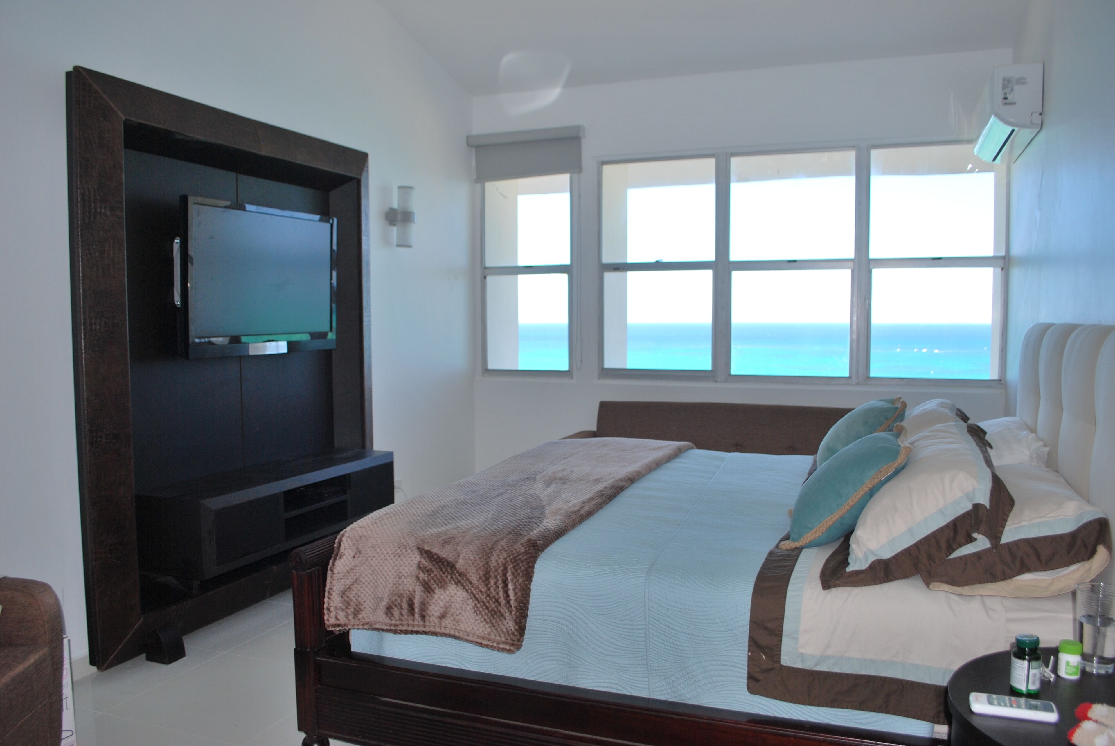 Un dormitorio con cama, televisor montado en un mueble de madera, ventana con vista al océano y mesita de noche con control remoto.