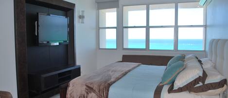 Un dormitorio con cama, televisor montado en un mueble de madera, ventana con vista al océano y mesita de noche con control remoto.
