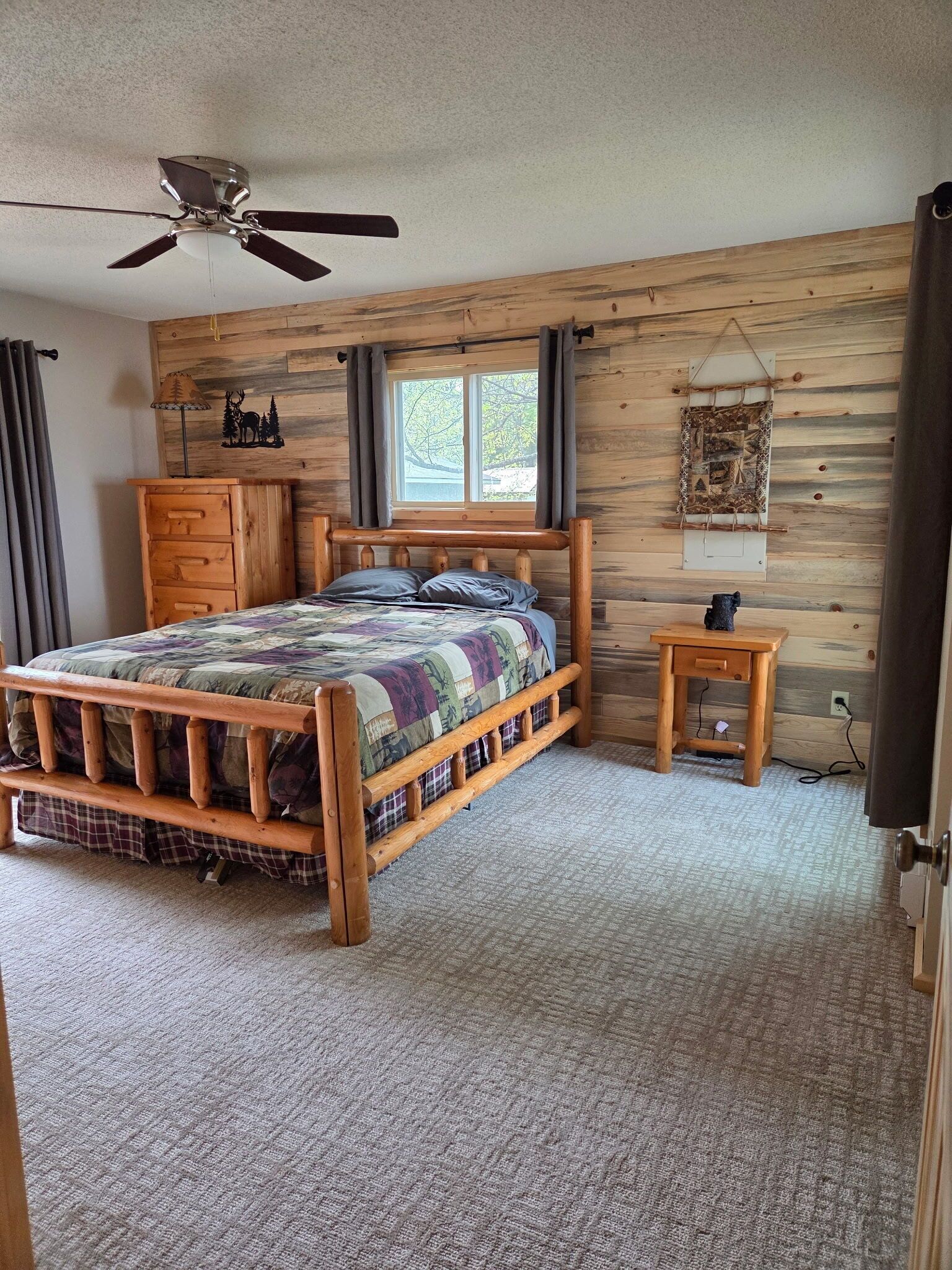 A wooden bedroom with a bed, a nightstand, a ceiling fan, a window, and a wall-mounted artwork.