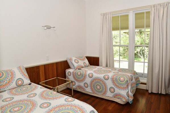 A room with two beds, a wooden headboard, a window with curtains, and a small table. - Fernridge Farm Cottage (Bellingen)