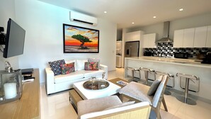 Ein modernes Wohnzimmer mit einem weißen Sofa, einem Couchtisch und einem Fernseher. Es gibt eine Küchenzeile mit weißen Schränken und einer gefliesten Spritzschutzwand.