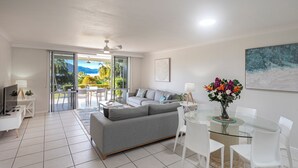 Una sala de estar moderna con un sofá gris, una mesa de comedor de vidrio, sillas blancas, un televisor y vistas a un jardín y montañas a través de amplios ventanales correderas.