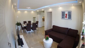A modern living room with a brown sofa, a dining area with chairs and a table, and a kitchen in the background.
