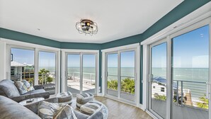 Ein geräumiges Wohnzimmer mit einem grauen Ecksofa, einem runden Couchtisch und einem Deckenventilator. Große bodentiefe Fenster bieten Blick auf den Ozean und eine Terrassenfläche.
