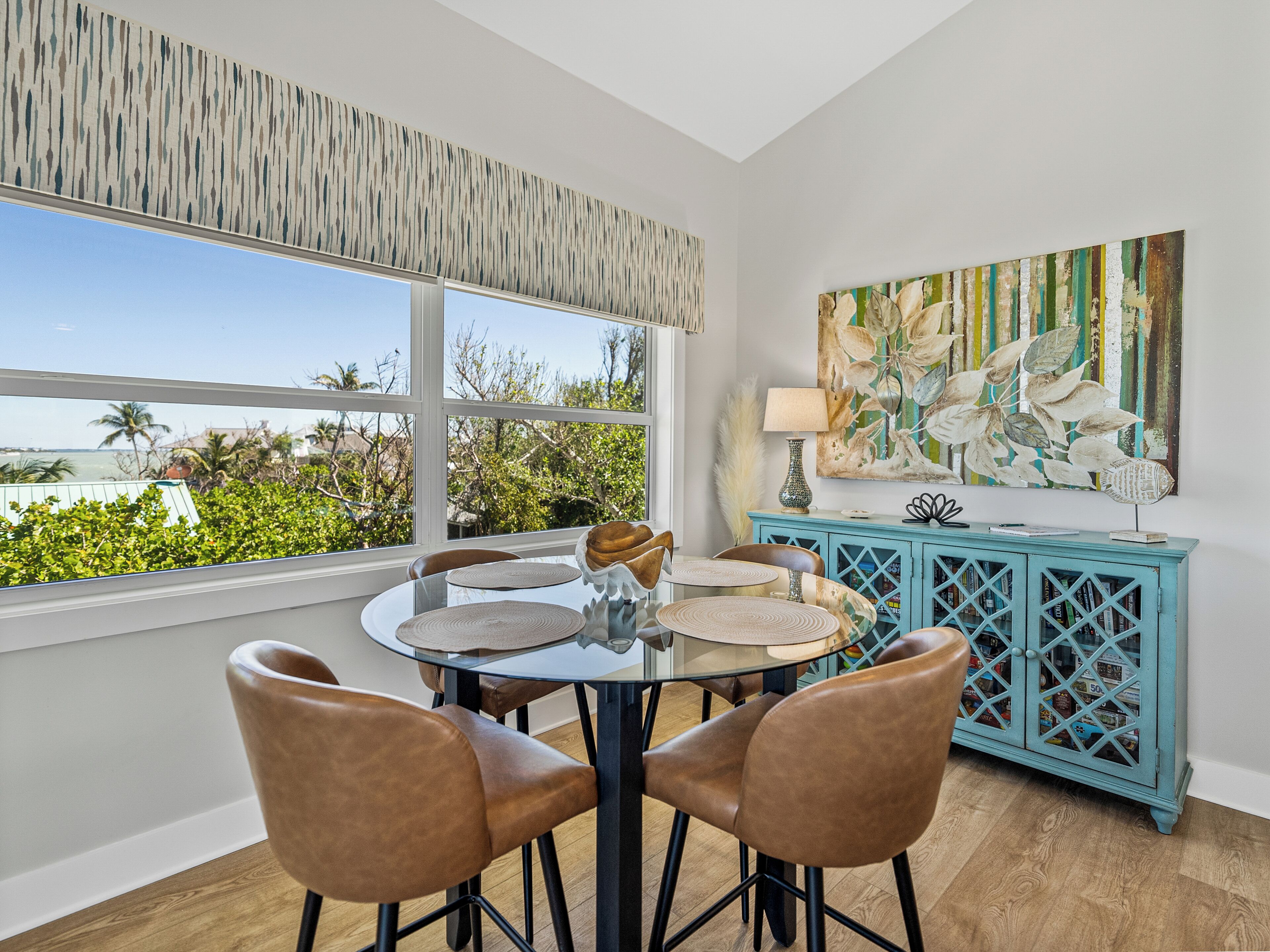 A dining area with a round table and four chairs, a teal cabinet with glass doors, and a large abstract painting on the wall.