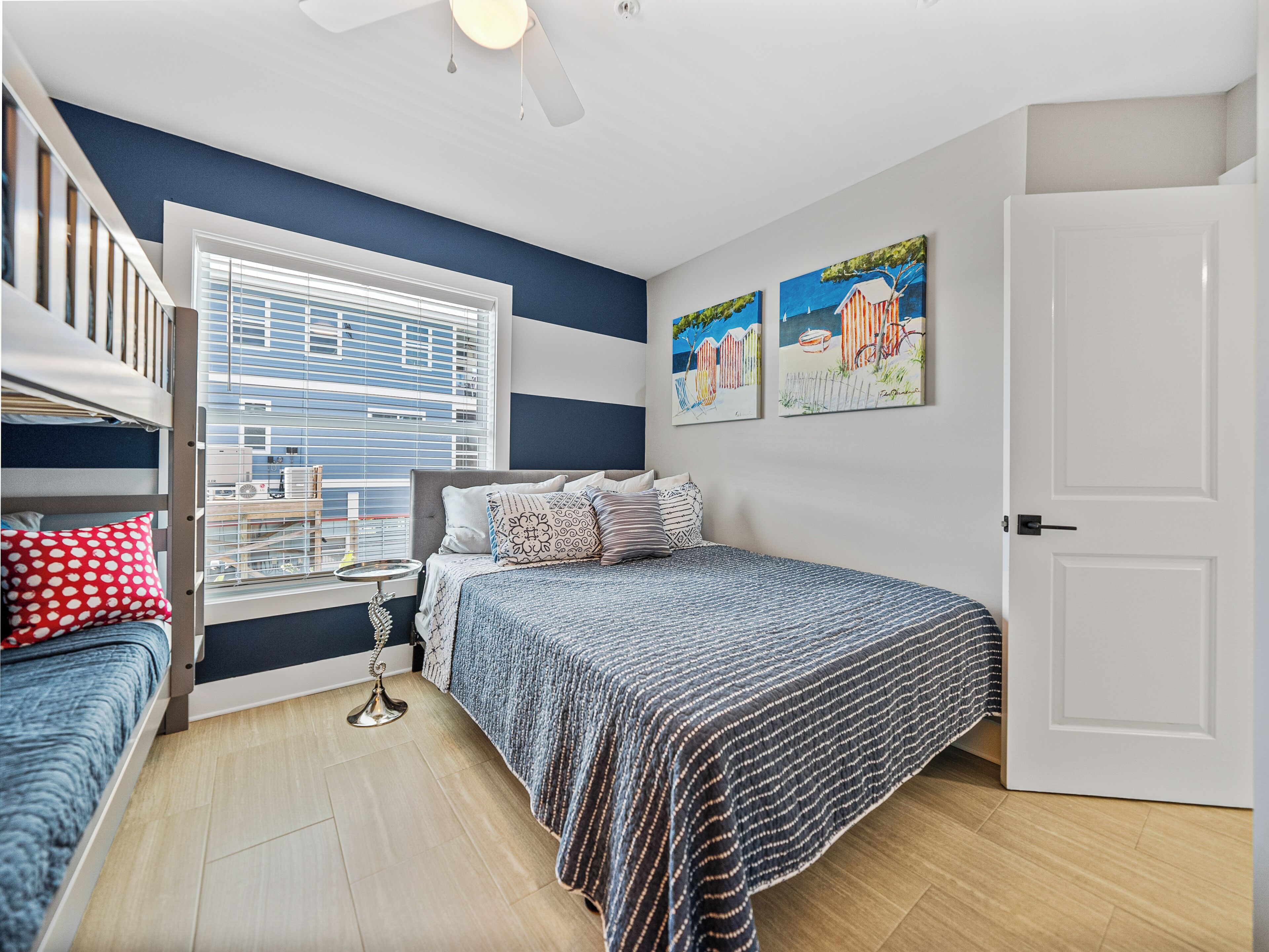 A bedroom with a bunk bed, a single bed, a window with blinds, a ceiling fan, and two paintings on the wall.