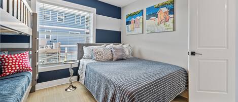 A bedroom with a bunk bed, a single bed, a window with blinds, a ceiling fan, and two paintings on the wall.