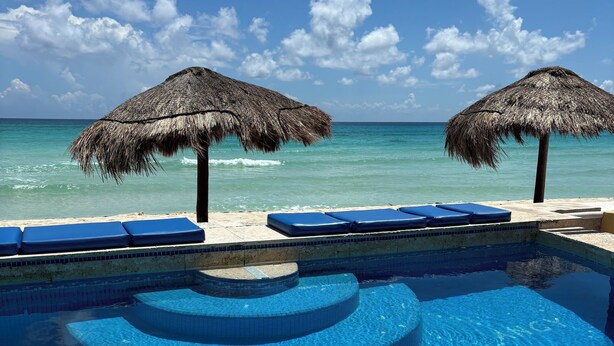 Una piscina con escalones de azulejos azules, dos sombrillas de paja y sillones de descanso junto a la piscina.