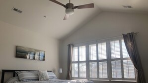 Ein Schlafzimmer mit Bett, Deckenventilator, Fenster mit Vorhängen und einem an der Wand befestigten Kunstwerk.