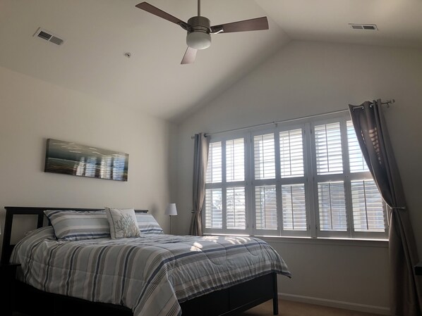 A bedroom with a bed, a ceiling fan, a window with curtains, and a wall-mounted artwork. - The Perfect Condo for Your Family! (Rehoboth Beach)