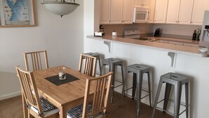 Ein Essbereich mit Holztisch und vier Stühlen, eine Küche mit weißen Schränken und einer Mikrowelle sowie eine Pendelleuchte.