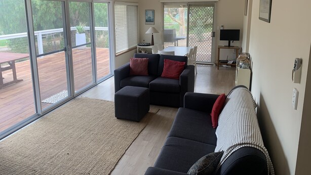 A spacious living room with a large sliding glass door leading to an outdoor deck, a dark-colored sofa with red cushions, a small table with a lamp, and a ceiling fan. - Port Willunga House on the Fleurieu Peninsula (Port Willunga)
