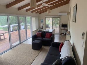 Un vaste salon doté d’une grande porte-fenêtre coulissante donnant sur une terrasse extérieure, d’un canapé de couleur sombre agrémenté de coussins rouges, d’une petite table avec une lampe et d’un ventilateur de plafond.