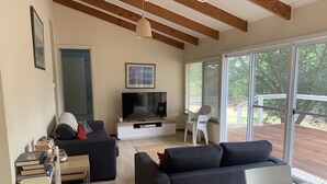 A modern living room with a flat-screen TV, dark sofas, a small table, and a large window offering a view of the outdoors. - Port Willunga House on the Fleurieu Peninsula (Port Willunga)