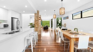 A modern kitchen with a central island, a dining area with a wooden table and chairs, and a view of greenery through a large window. - Luxury Townhouse 150 metres to the beach (Torquay)