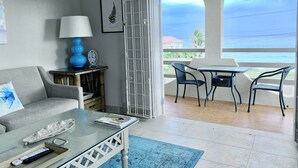 A living room with a sofa, a coffee table, a lamp, and a view of the ocean through an open door.