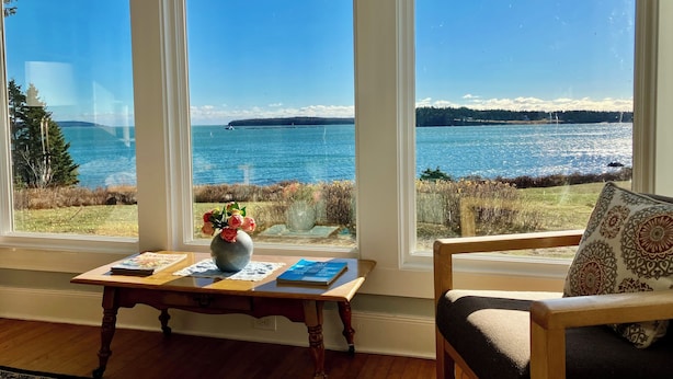 Ein Zimmer mit großem Fenster mit Blick auf ein Gewässer, einem Holztisch mit Büchern und einem Blumenstrauß sowie einem Sessel mit gemustertem Kissen.
