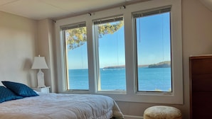 Ein Schlafzimmer mit einem großen Bett, einem Nachttisch mit Lampe, einem kleinen Hocker und Blick auf einen See durch drei Fenster.