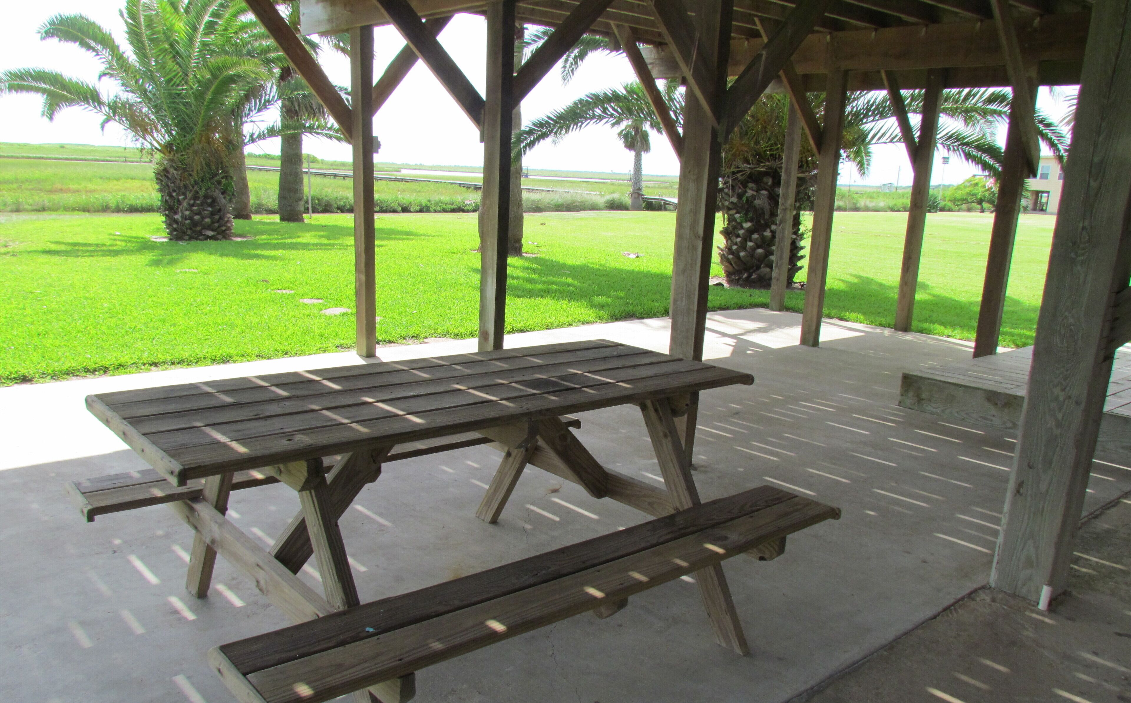 Una mesa de picnic de madera con bancos bajo un pabellón de madera con suelo de concreto, rodeado de un área cubierta de césped con palmeras.