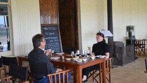 Ein Restaurant mit Holztischen und -stühlen, einer Kreidetafel als Speisekarte und Blick ins Freie.