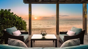 Un espacio de estar techado al aire libre con muebles de mimbre y una mesa, con vista a una playa al atardecer.
