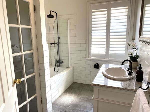 A bathroom with a glass-enclosed shower, a white bathtub, a sink with a marble countertop, and a window with blinds.