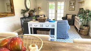 Una sala de estar con una consola blanca, un sofá azul, una mesa de comedor con frutero y un ventanal con vista a follaje.