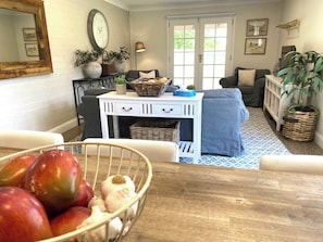 A living room with a white console table, a blue sofa, a dining table with a fruit bowl, and a large window with a view of greenery.
