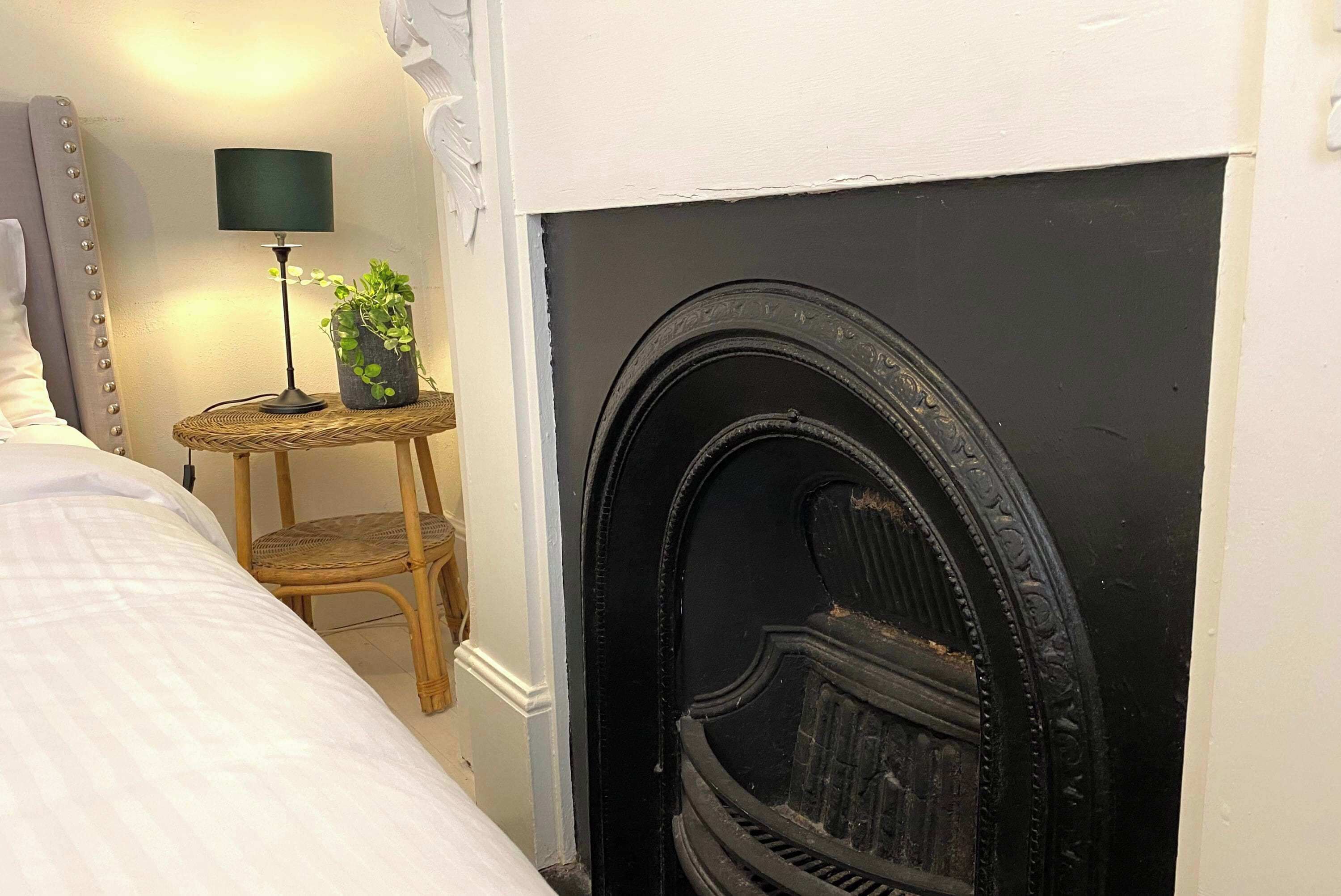 A bedroom with a bed, a small table with a lamp, a chair, and a fireplace.