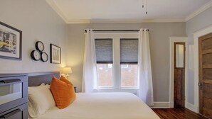 Ein Schlafzimmer mit einem Bett, einem Nachttisch, einer Lampe, einem Fenster mit Vorhängen, einer Tür und einer Wanduhr.