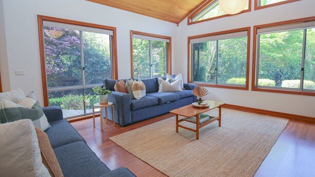 A spacious living room with a blue sofa, a wooden coffee table, and large windows offering a view of greenery. - ON THE TRAIL : Bright (Bright)