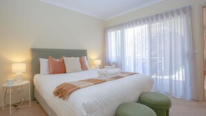 A hotel room with a large bed, two bedside lamps, a nightstand, a green ottoman, and a window with sheer curtains. - ON THE TRAIL : Bright (Bright)
