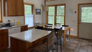 A kitchen with wooden cabinets, a large island, and a dining area with chairs. - Tranquil Views of Drummer Cove (1321) (Wellfleet)