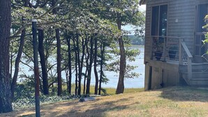 A lakeside cabin with a deck overlooking the water, surrounded by tall trees. - Tranquil Views of Drummer Cove (1321) (Wellfleet)