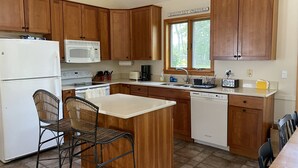A kitchen with wooden cabinets, a white refrigerator, a microwave, a stove, a sink, and a window with a view of trees. - Tranquil Views of Drummer Cove (1321) (Wellfleet)
