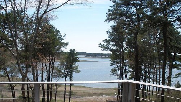A wooden deck with a metal railing overlooking a lake, surrounded by trees. - Tranquil Views of Drummer Cove (1321) (Wellfleet)