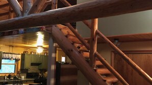 Un interior rústico con una escalera de madera, una zona de estar acogedora y una cocina visible al fondo.