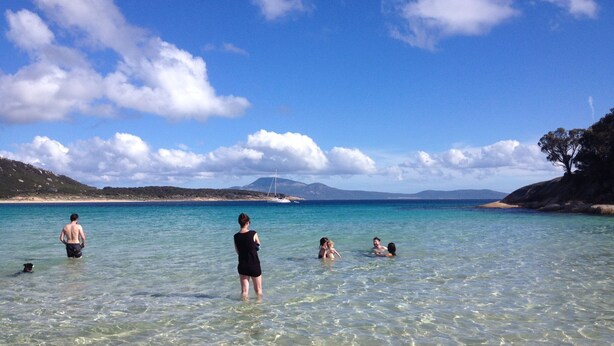 Ein Strand mit klarem türkisblauem Wasser, Menschen, die schwimmen und im Wasser stehen, und ein Segelboot in der Ferne.