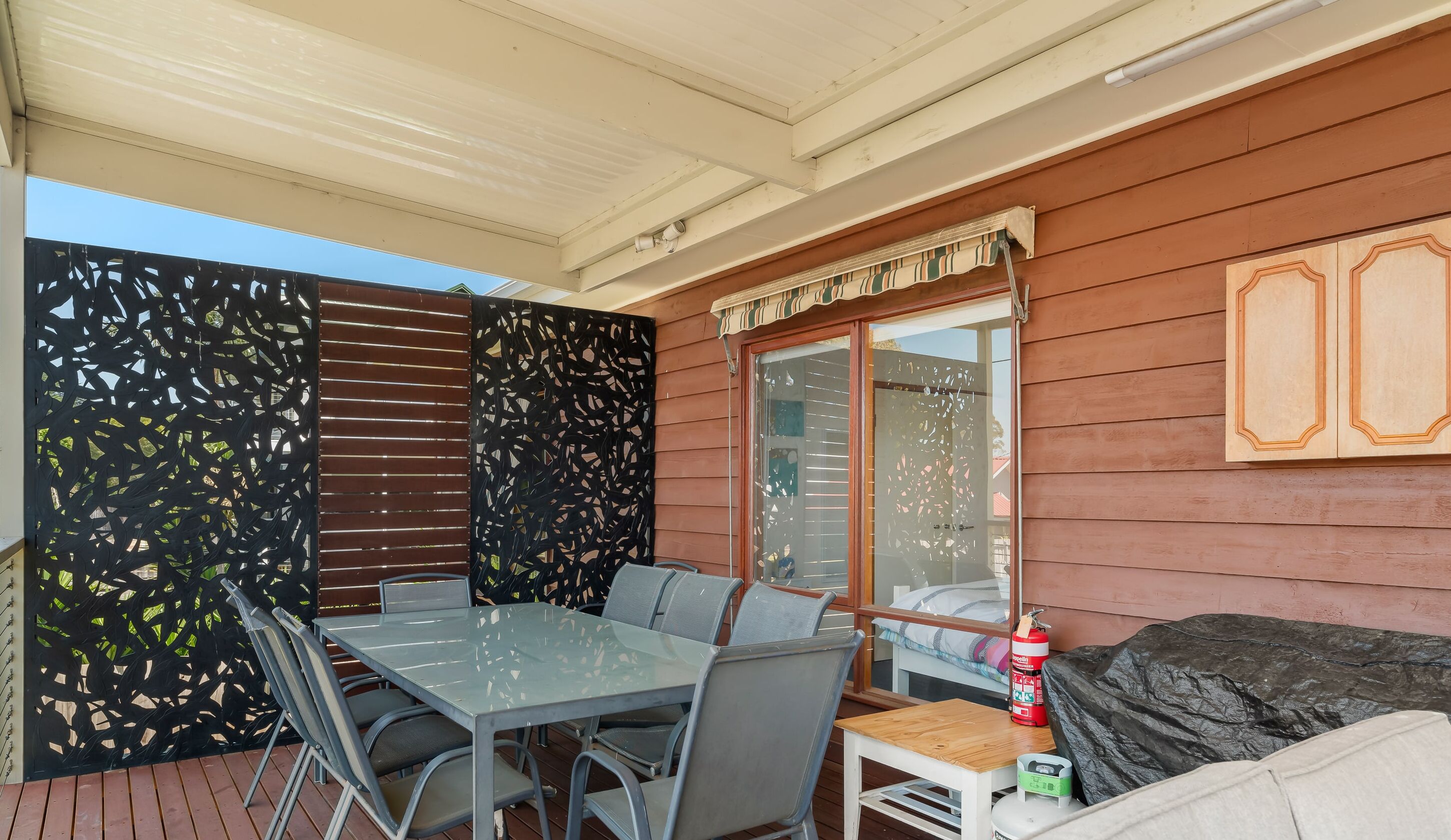 A covered outdoor seating area with a table and chairs, a barbecue grill, and a fire extinguisher.