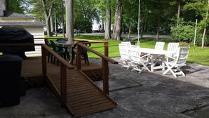 Una terraza de madera con una mesa, sillas, una parrilla y vistas a un camino y árboles.