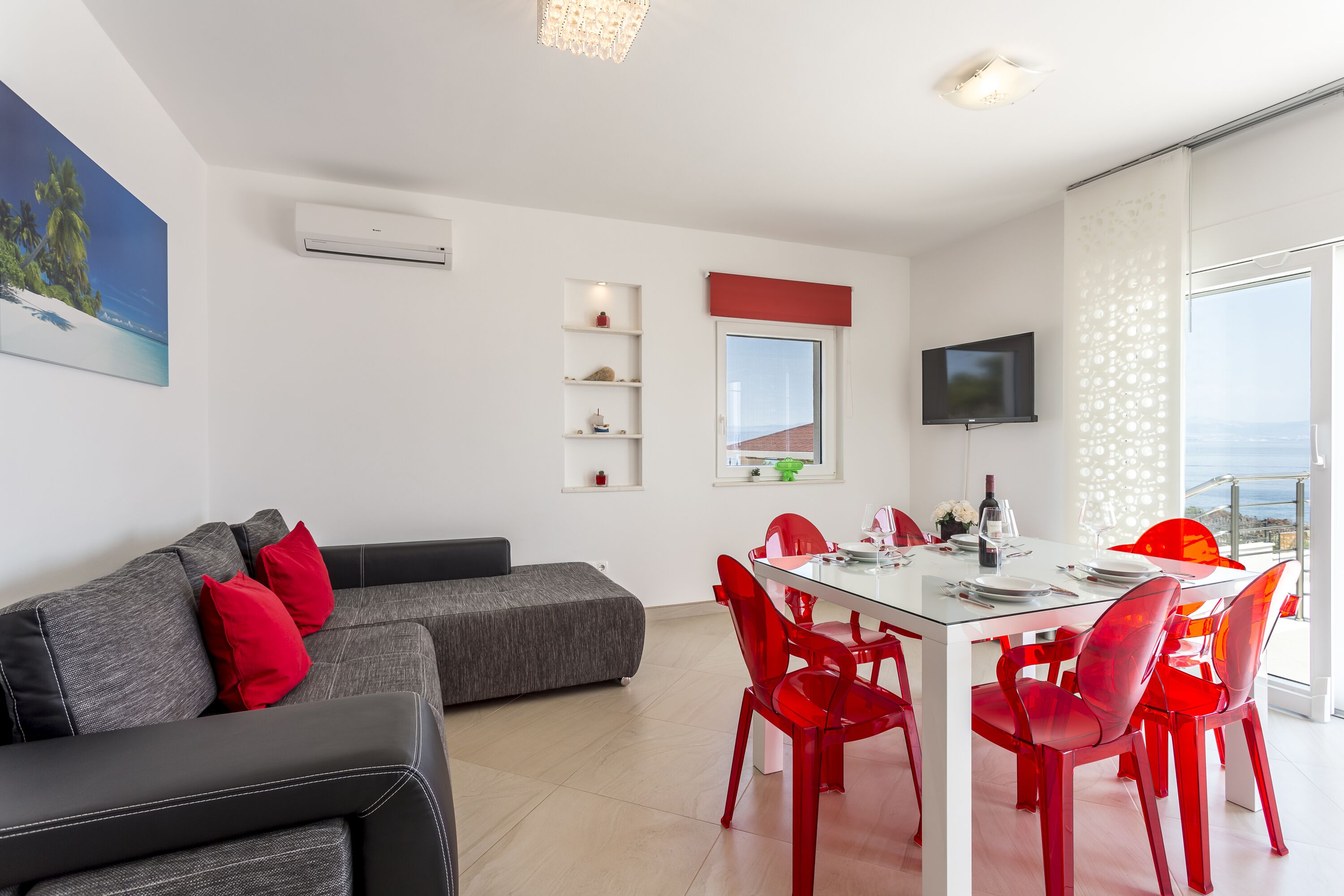 Ein modernes Wohnzimmer mit grauem Sofa, roten Kissen, einem Glastisch mit roten Stühlen, einem Flachbildfernseher und einem großen Fenster mit Blick.