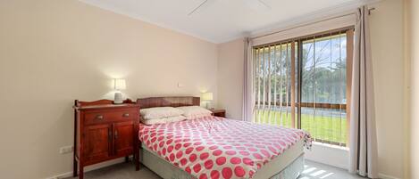 A bedroom with a bed, wooden dresser, two bedside lamps, a ceiling fan, and a large window with curtains.