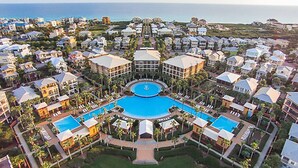 An aerial view of a resort with multiple buildings, a central swimming pool, and a view of the ocean.