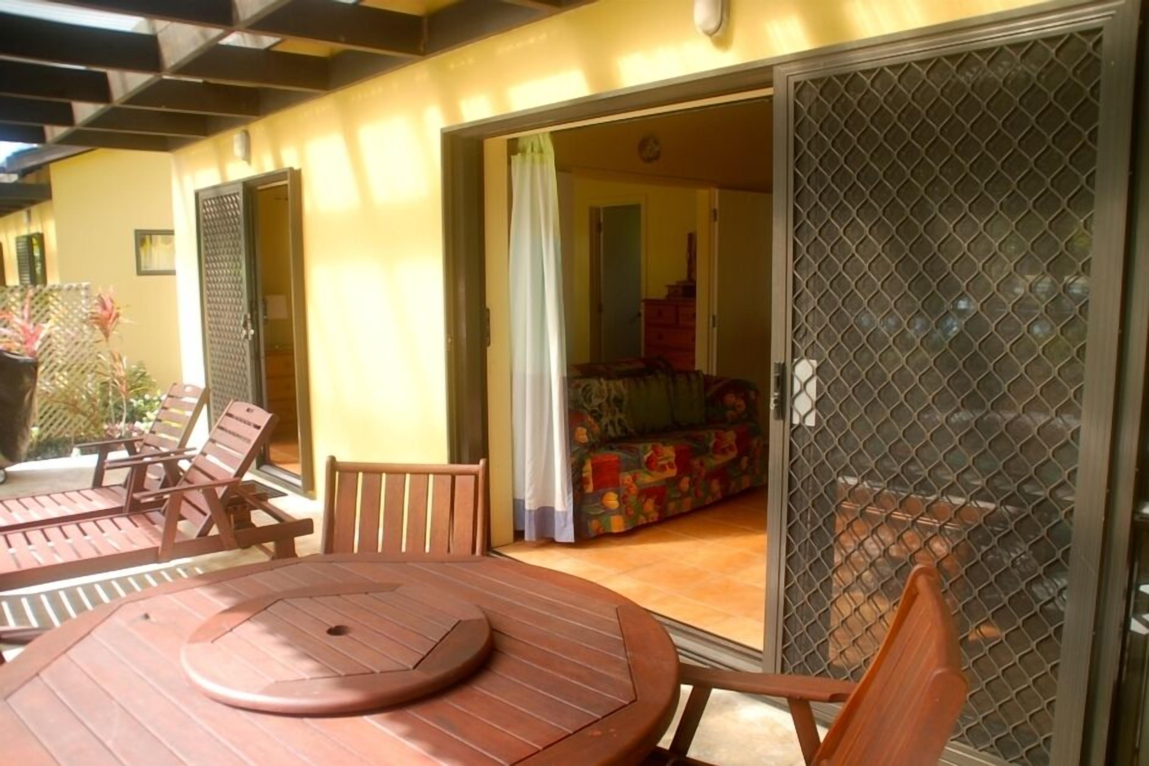 Un patio ensoleillé avec une table et des chaises en bois, une porte-fenêtre coulissante donnant sur une pièce où se trouvent un canapé coloré et un lit.