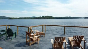 Une terrasse dotée de chaises et de tables en bois, offrant une vue sur un lac paisible dont les rives sont bordées d’une forêt.
