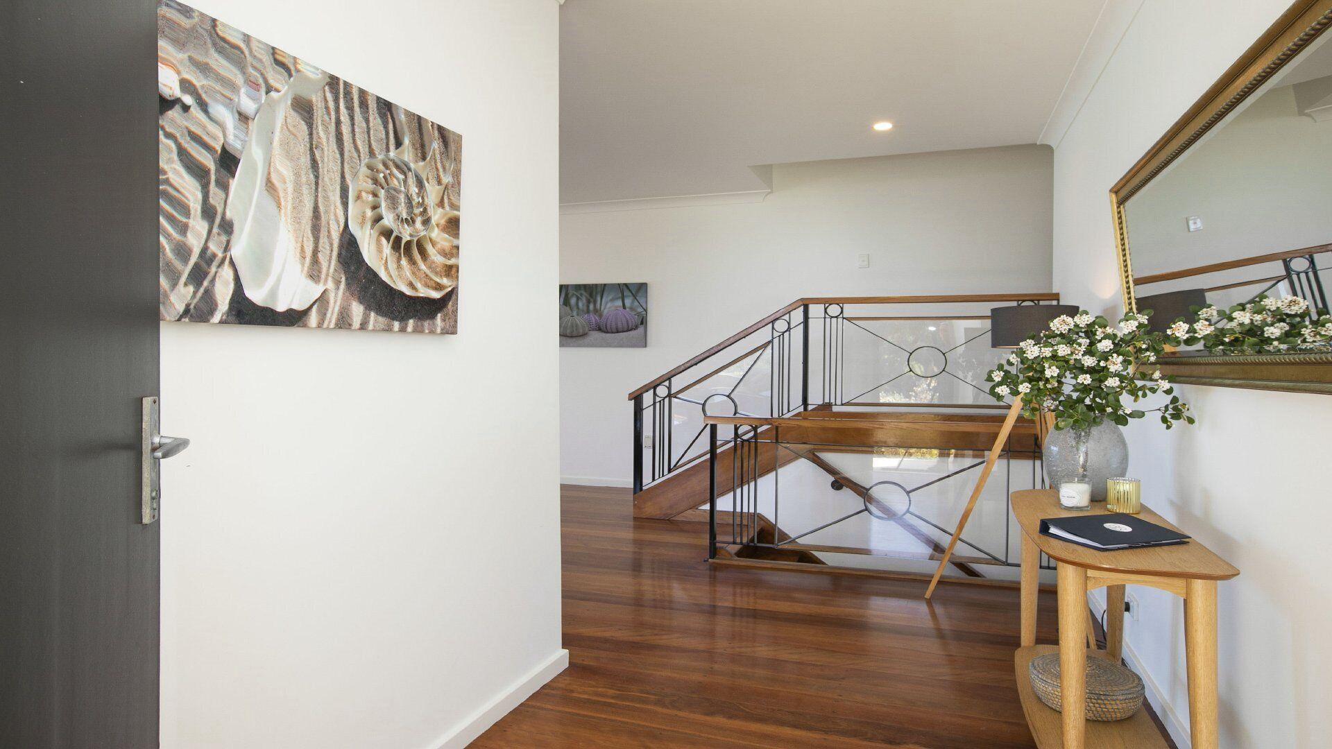A hallway with a wooden staircase, a door with a metal handle, a framed artwork, and a small table with a vase and a candle.